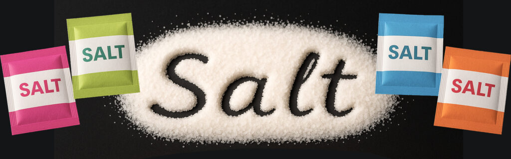 The word "Salt" written in a bed of coarse salt granules on a black surface, surrounded by colorful single-serve salt sachets in pink, green, blue, and orange.