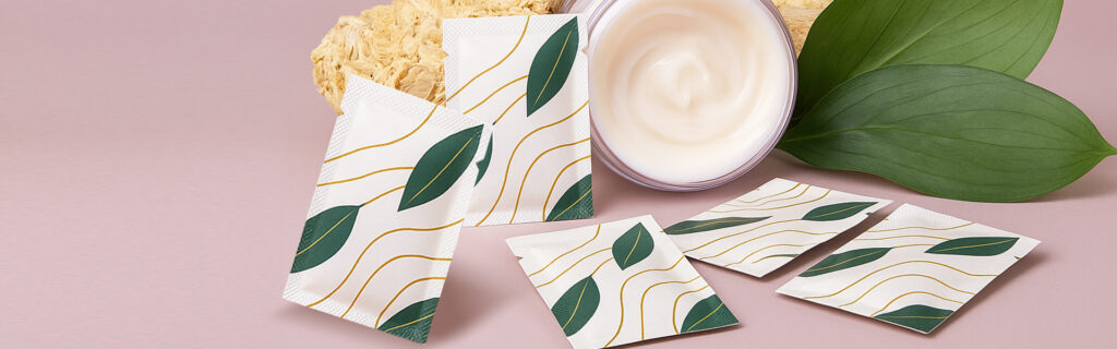 Small cosmetic cream sachets with a clean, botanical design arranged beside an open jar of cream and green leaves on a soft pink background.