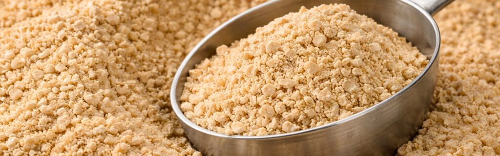 Close-up of granular medicated feed additive in a stainless-steel scoop surrounded by bulk product.