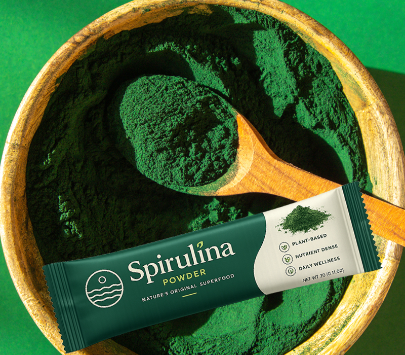Spirulina packaging in stick packs displayed beside green spirulina powder and a wooden scoop.